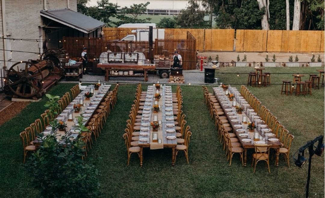 Three long banquet tables on the lawn at dusk, a chef’s station and bar behind, festoon lights overhead