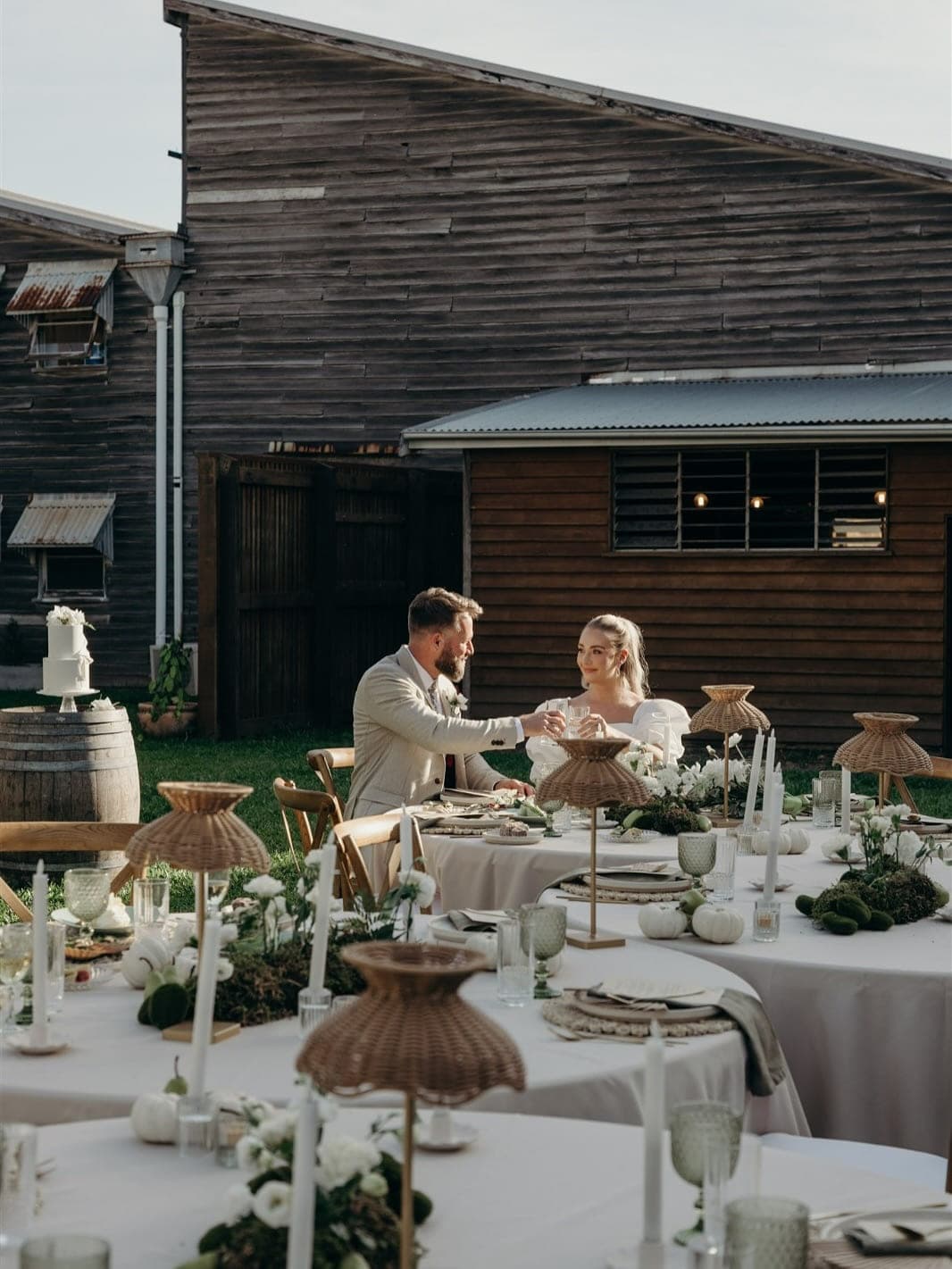 A couple toasting at a round table on the lawn at golden hour, the mill’s timber sheds behind, wicker lamps and white linen
