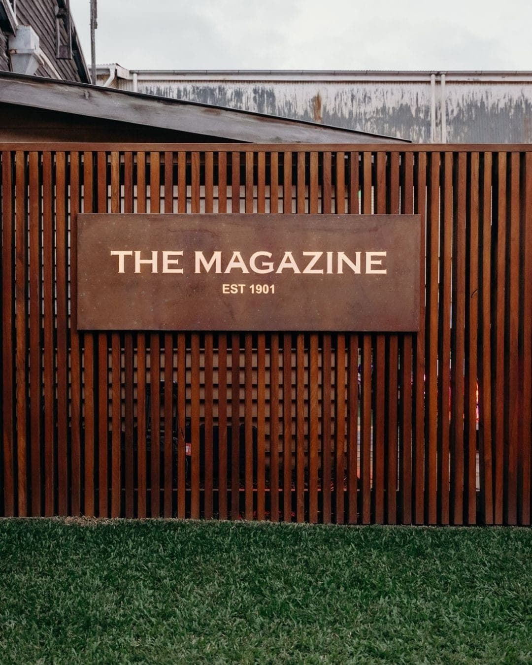 Corten steel sign reading The Magazine, est. 1901, mounted on a timber batten screen above the lawn