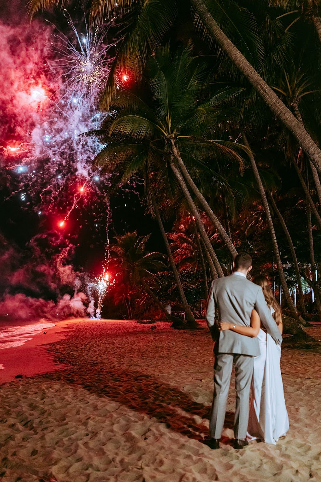 The same couple on the sand as red and pink fireworks light up the palms and the smoke drifts over the water
