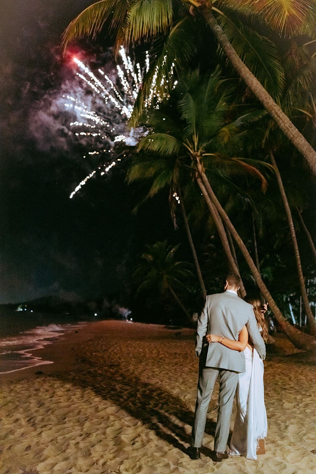A bride and groom seen from behind, arms around each other on the beach at night, white fireworks bursting above the palms