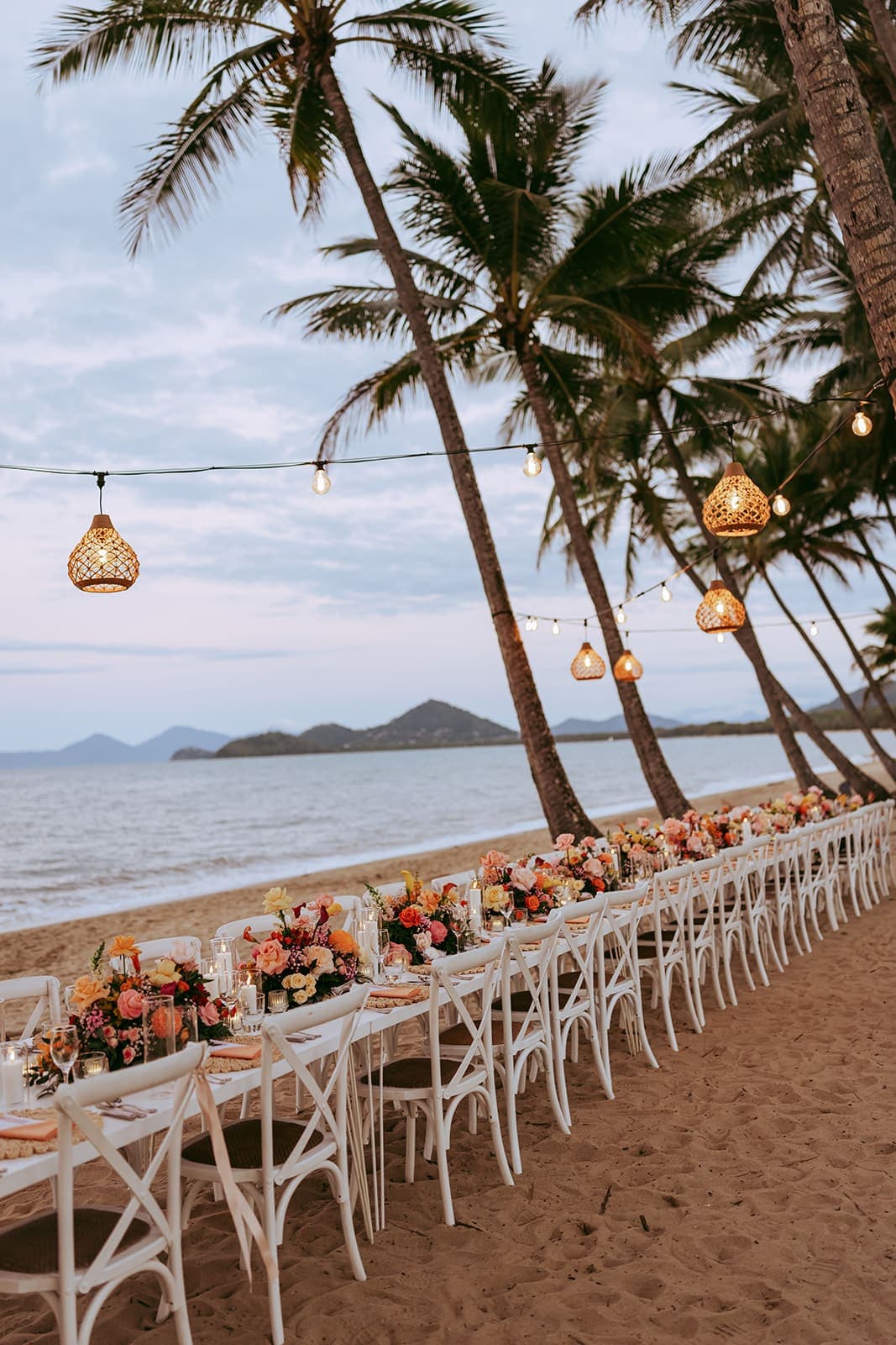 A long wedding table on the sand at dusk, white cross-back chairs and bright flowers down the middle, lanterns strung through leaning palms, hills across the water