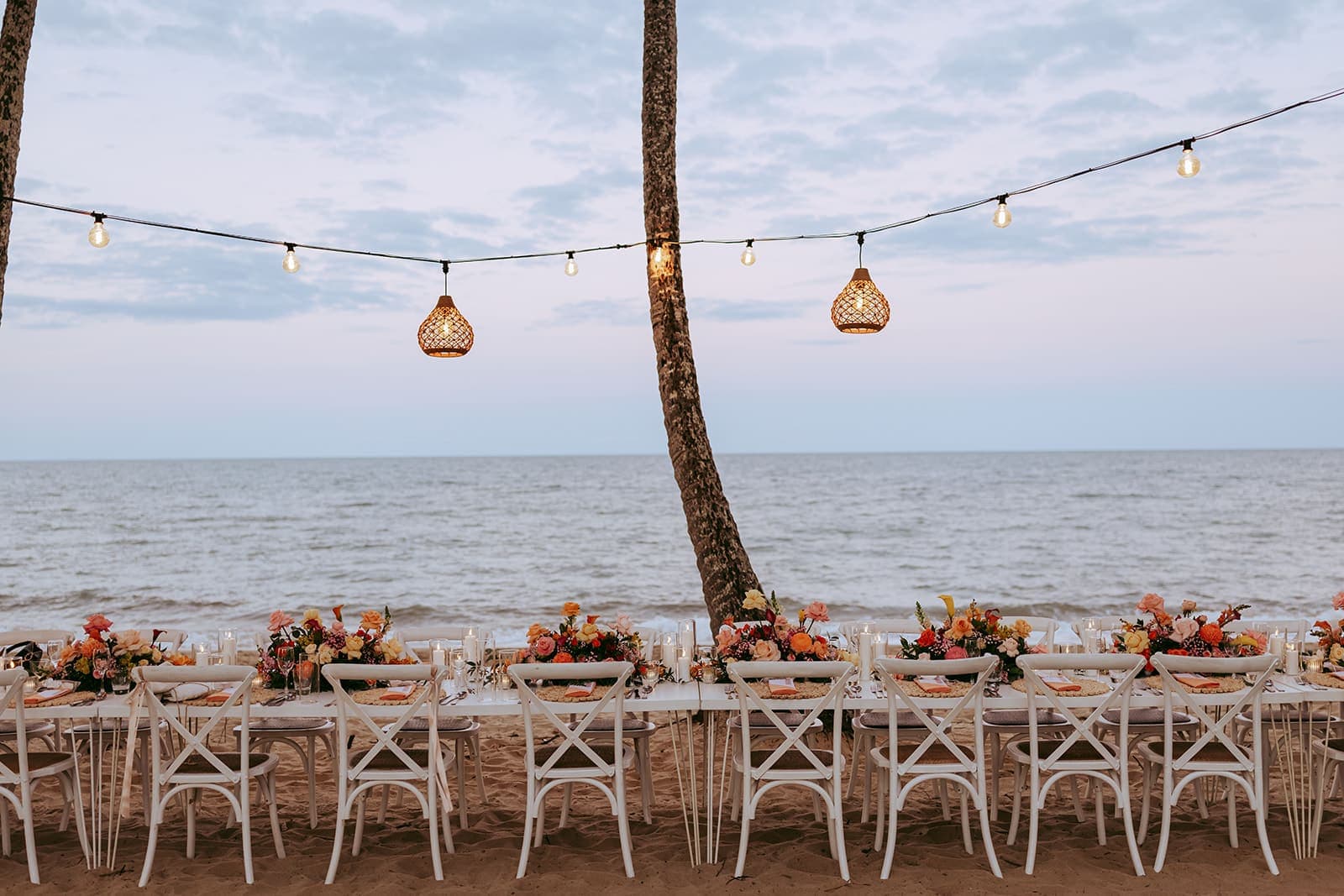A wedding table set on the sand, white cross-back chairs, candles and orange and pink flowers, two woven lanterns hanging over it and calm sea behind