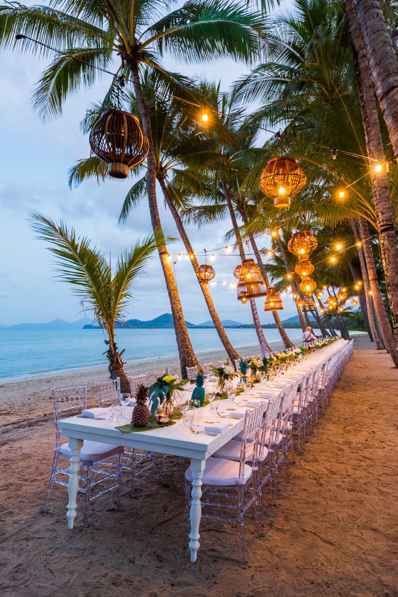 A long white table set for dinner on the sand at Palm Cove, lanterns strung through the palms